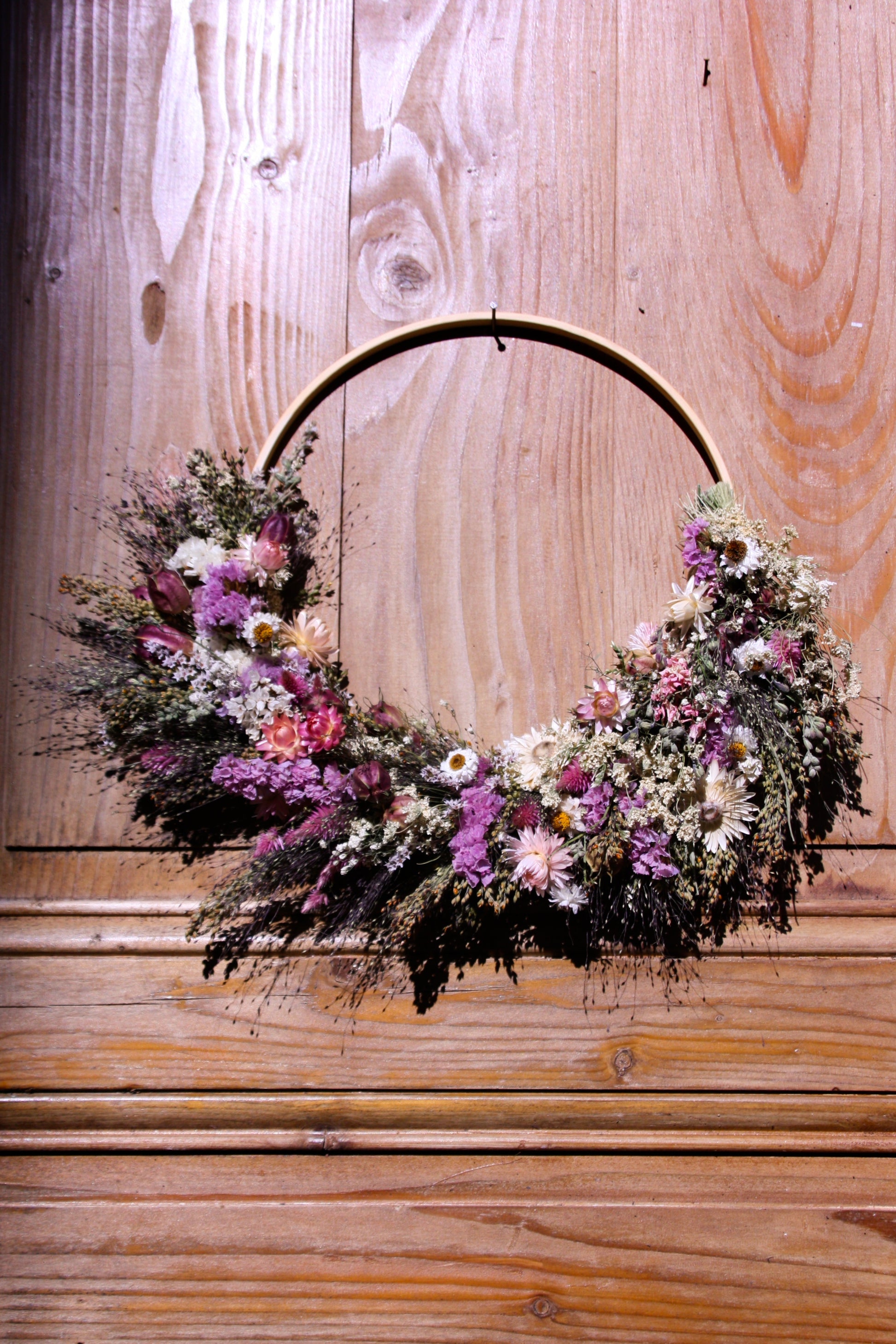 Couronne murale en fleurs séchées naturelles tressée en demi-lune sur un cercle en bois. Création artisanale aux tons roses et blancs